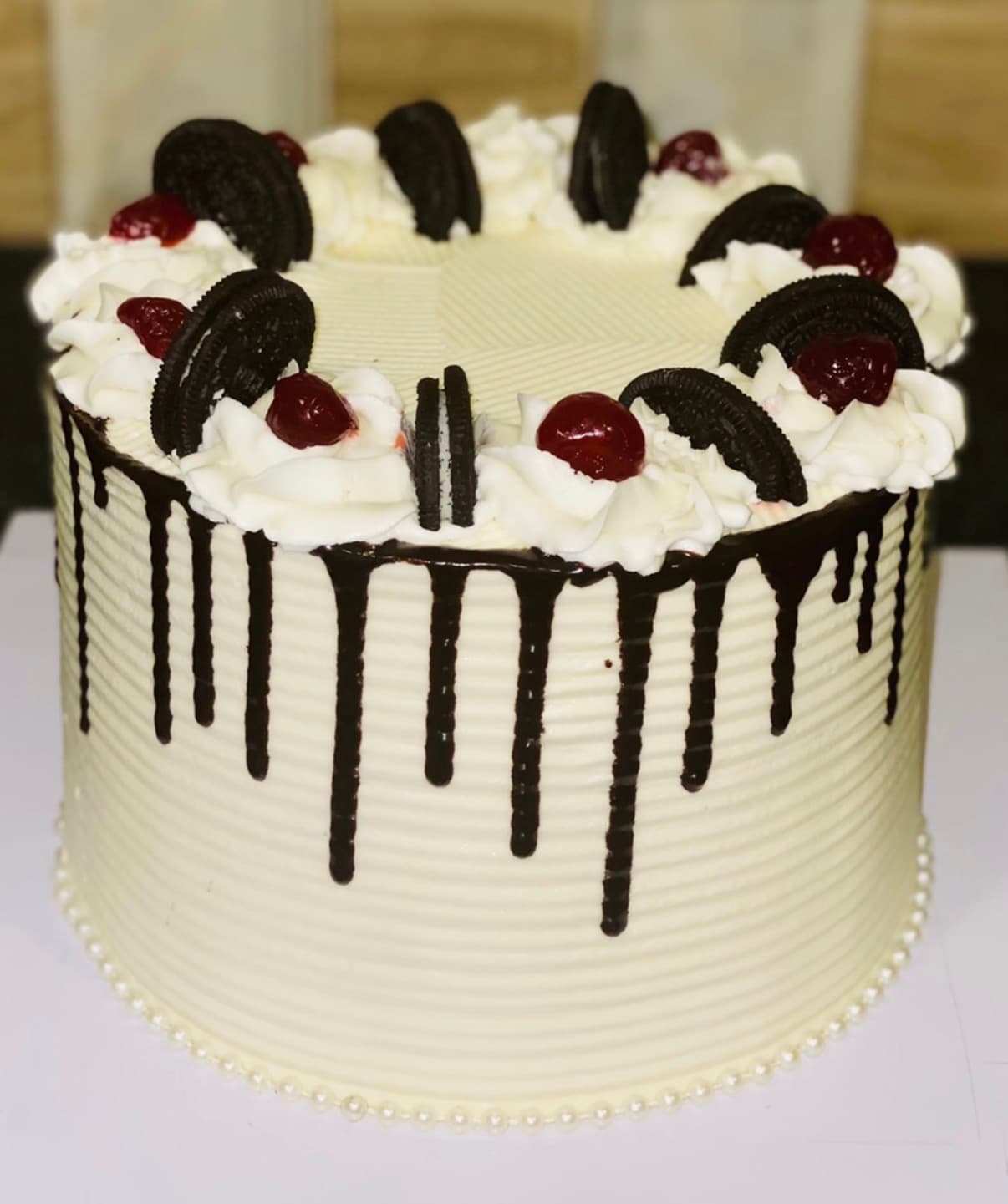 Ivory celebration cake with dark drip styling, sandwich cookies, cream swirls, and cherries